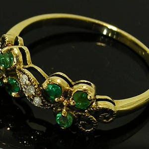 Op de afbeelding: Een gouden ring met een bloemmotief, bezet met smaragdgroene edelstenen en kleine witte diamanten. De ring heeft een delicate band en gedetailleerde zetting rond de edelstenen.