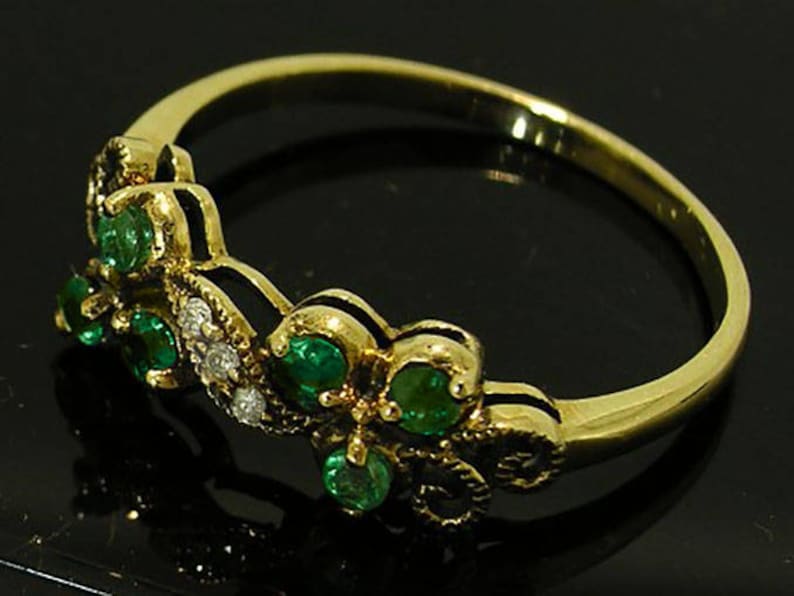 Op de afbeelding: Een gouden ring met een bloemmotief, bezet met smaragdgroene edelstenen en kleine witte diamanten. De ring heeft een delicate band en gedetailleerde zetting rond de edelstenen.