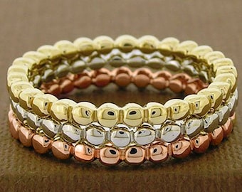 Op de afbeelding: Drie gestapelde ringen in goud, zilver en roségoud. Elke ring heeft een kralenontwerp, waardoor een textuur ontstaat. De ringen worden gepresenteerd op een bruin oppervlak, dat het contrast van kleuren en texturen laat zien.