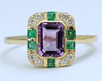 Op de afbeelding: Een gouden ring met een rechthoekige, lichtpaarse amethist middensteen. De ring is geaccentueerd met kleine, ronde witte diamanten en vierkante groene smaragden. De ring heeft een klassiek art deco-ontwerp.