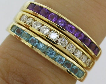Op de afbeelding: Drie gouden ringen op elkaar gestapeld, elk met een rij edelstenen. De bovenste ring heeft paarse amethiststenen, de middelste ring heeft heldere diamanten en de onderste ring heeft blauwe aquamarijnstenen. De ringen zijn gezet in een geelgouden band.