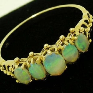 May include: A gold ring featuring five oval opals. The opals have a play of color, with hints of green and blue. The ring's band and setting are ornate, with a detailed design. The ring is set against a dark background.