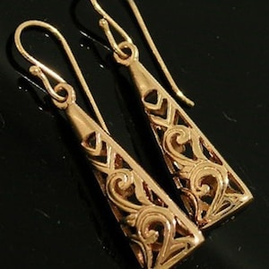 May include: A pair of gold-colored, openwork earrings. Each earring has a long, triangular shape with intricate cut-out designs and a hook closure. The earrings are displayed against a dark background.