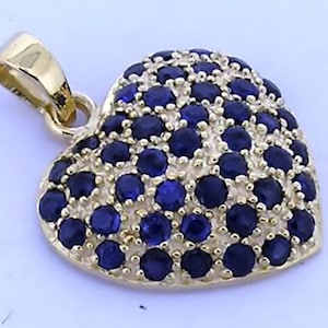 Puede incluir: Un colgante en forma de corazón de color dorado con una argolla. El colgante está incrustado con numerosas piedras preciosas pequeñas, redondas y de color azul oscuro. Las piedras preciosas cubren toda la superficie frontal del corazón, creando un efecto brillante. La argolla también es dorada.