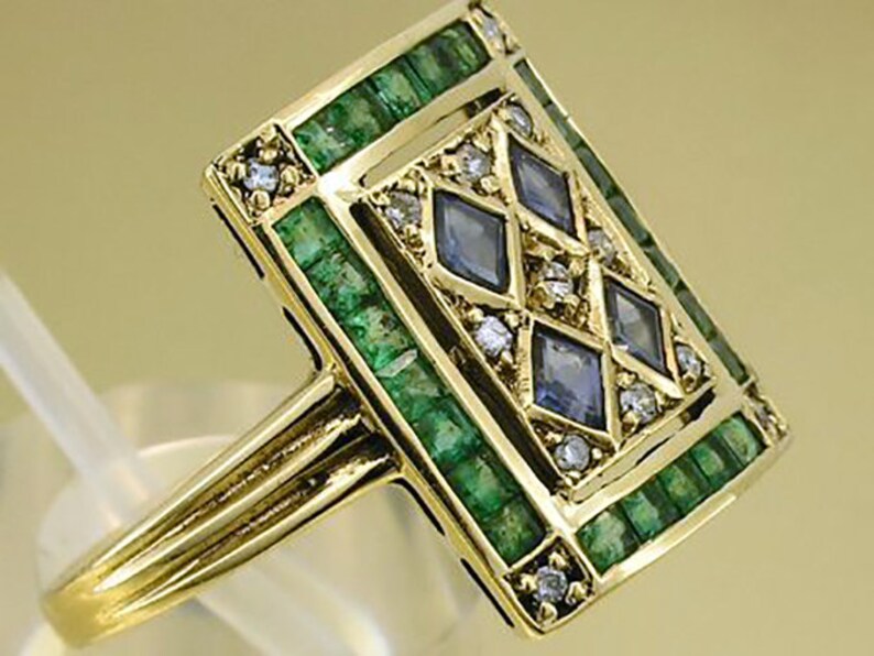 Pu&ograve; includere: Anello in oro con una faccia rettangolare decorata con smeraldi, pietre preziose blu e piccoli diamanti. Il gambo dell'anello &egrave; diviso in tre sezioni parallele. Gioiello di lusso.