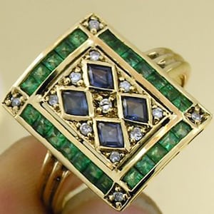 Pu&ograve; includere: Un anello d'oro con un design geometrico. L'anello presenta un quadrato centrale con quattro pietre quadrate blu, circondato da pietre verdi taglio smeraldo e piccoli diamanti. La fascia dell'anello &egrave; in oro lucido semplice.