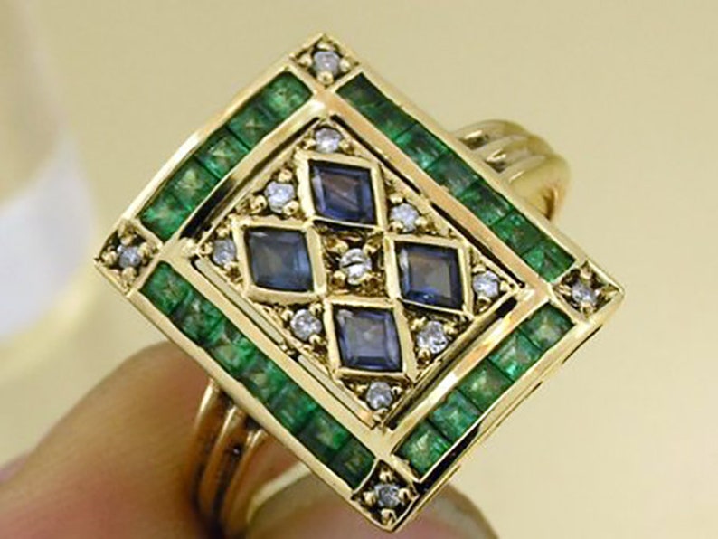 Pu&ograve; includere: Un anello d'oro con un design geometrico. L'anello presenta un quadrato centrale con quattro pietre quadrate blu, circondato da pietre verdi taglio smeraldo e piccoli diamanti. La fascia dell'anello &egrave; in oro lucido semplice.