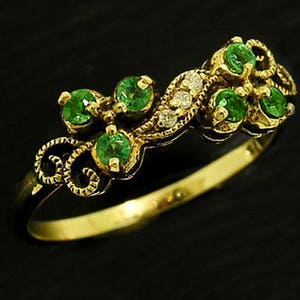 Op de afbeelding: Een gouden ring met een reeks ronde, heldergroene edelstenen en kleine diamanten. De ring heeft een sierlijk, wervelend ontwerp op de band en rond de edelstenen. De ring is geplaatst tegen een zwarte achtergrond.