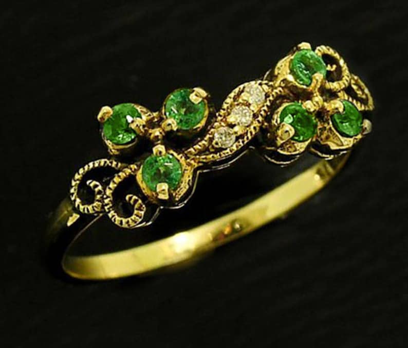 Op de afbeelding: Een gouden ring met een reeks ronde, heldergroene edelstenen en kleine diamanten. De ring heeft een sierlijk, wervelend ontwerp op de band en rond de edelstenen. De ring is geplaatst tegen een zwarte achtergrond.