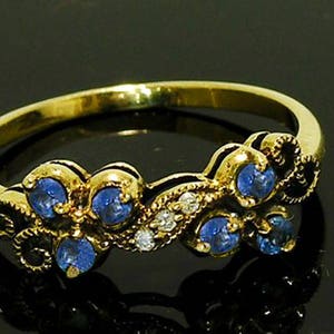 Op de afbeelding: Een gouden ring met een bloemmotief, bezet met blauwe edelstenen en kleine witte stenen. De ring heeft een gepolijste afwerking en wordt getoond tegen een donkere achtergrond. De edelstenen zijn rond en in een decoratief patroon geplaatst.