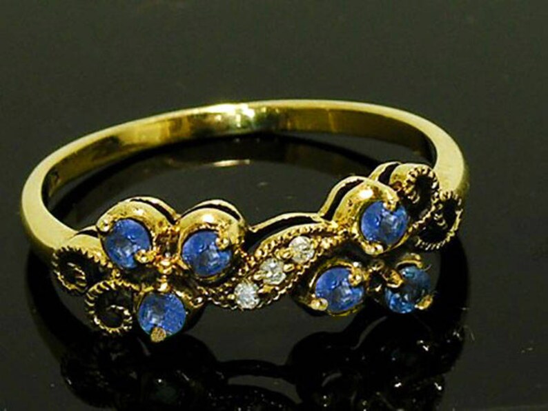 Op de afbeelding: Een gouden ring met een bloemmotief, bezet met blauwe edelstenen en kleine witte stenen. De ring heeft een gepolijste afwerking en wordt getoond tegen een donkere achtergrond. De edelstenen zijn rond en in een decoratief patroon geplaatst.