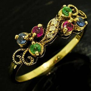 Op de afbeelding: Een gouden ring met een bloemmotief en een cluster van edelstenen. De ring heeft een gouden band en een decoratieve bovenkant met rode, groene, blauwe en heldere edelstenen. De ring staat tegen een donkere achtergrond.