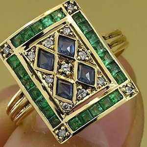 Pu&ograve; includere: Un anello d'oro con una faccia rettangolare, con un bordo di smeraldi verdi. Il design centrale include quattro gemme blu a forma di diamante, circondate da piccoli diamanti. L'anello ha un gambo a pi&ugrave; bande.