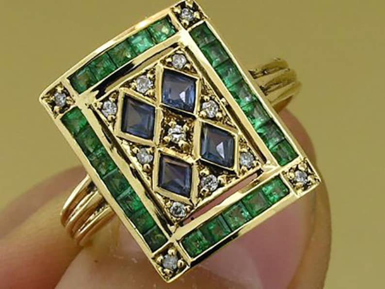 Pu&ograve; includere: Un anello d'oro con una faccia rettangolare, con un bordo di smeraldi verdi. Il design centrale include quattro gemme blu a forma di diamante, circondate da piccoli diamanti. L'anello ha un gambo a pi&ugrave; bande.