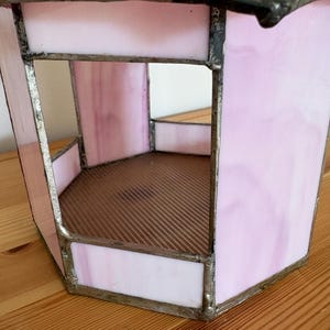 May include: A pink stained glass lantern with a hexagonal shape. The glass is a soft pink with darker pink marbling. The edges are framed with a silver-colored metal. The interior has a brown, corrugated base.