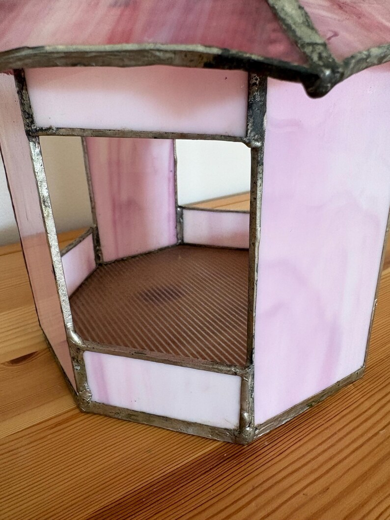 May include: A pink stained glass lantern with a hexagonal shape. The glass is a soft pink with darker pink marbling. The edges are framed with a silver-colored metal. The interior has a brown, corrugated base.