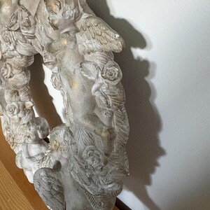 Op de afbeelding: Een gedetailleerde, gebroken witte, cirkelvormige krans met cherubijnen met vleugels en rozen. De krans heeft een verweerde afwerking met gouden accenten, wat een vintage esthetiek cre&euml;ert. De krans is gemaakt van een stevig materiaal en is geschikt voor wanddecoratie.
