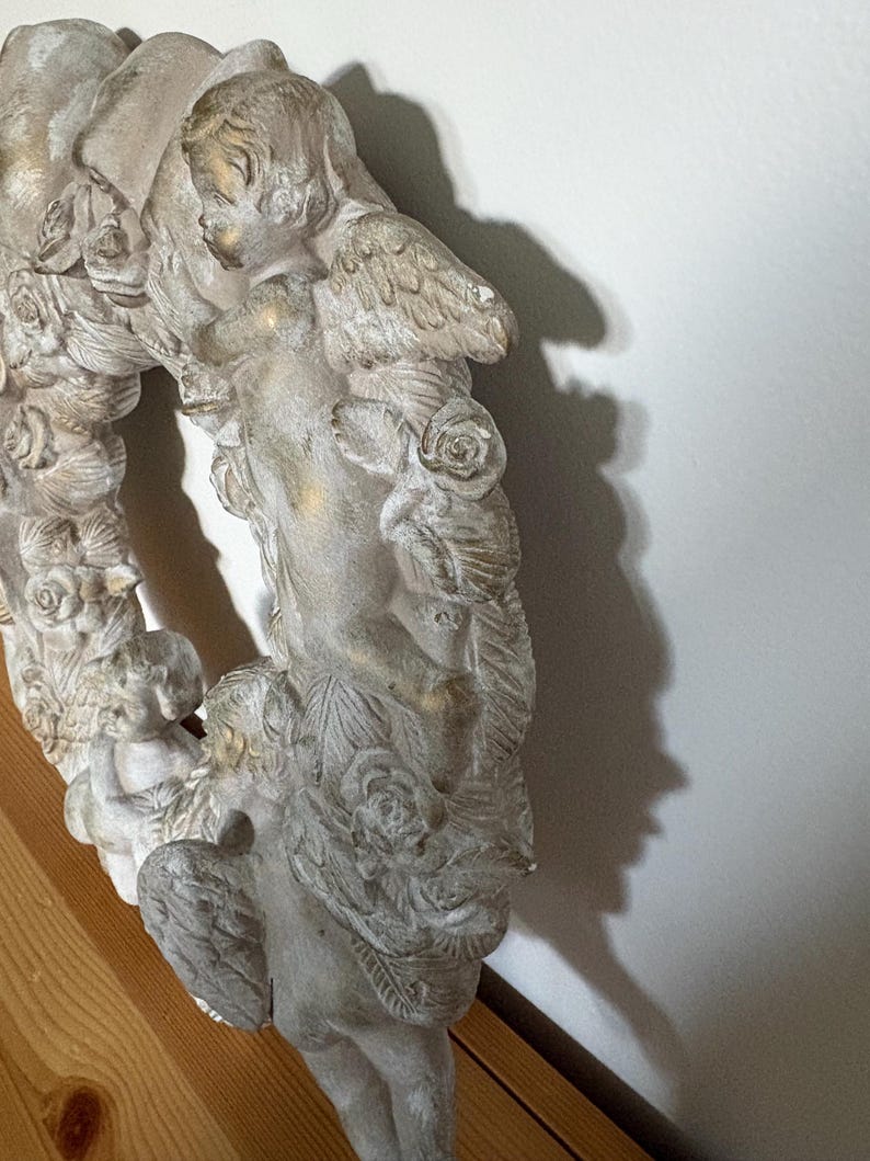 Op de afbeelding: Een gedetailleerde, gebroken witte, cirkelvormige krans met cherubijnen met vleugels en rozen. De krans heeft een verweerde afwerking met gouden accenten, wat een vintage esthetiek cre&euml;ert. De krans is gemaakt van een stevig materiaal en is geschikt voor wanddecoratie.