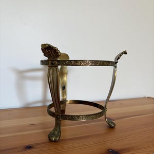Könnte beinhalten: Ein goldfarbener dekorativer Metallständer mit runder Oberseite und drei Beinen. Die Beine sind wie Löwenpfoten geformt und durch einen dekorativen Ring verbunden. Der Ständer hat ein verziertes Design.