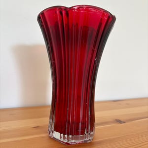 May include: A ruby red glass vase with a fluted design. The vase has a square base and a flared, scalloped top. The vertical fluting creates a textured effect, enhancing the rich color. The vase is suitable for displaying flowers or as a decorative piece.