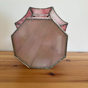 May include: A vintage, octagonal stained glass lamp shade. The shade features a pink and red color scheme with a ribbed texture on the main body. The edges are trimmed with a silver metal. The lamp shade is sitting on a wooden surface.