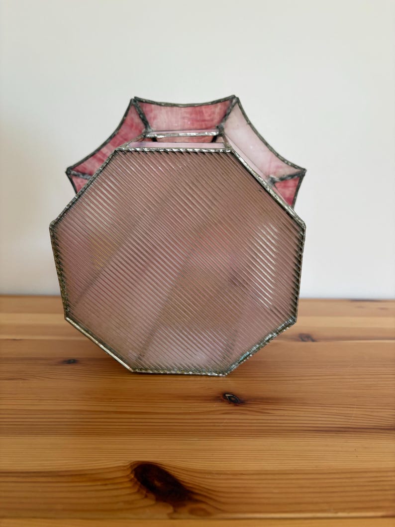 May include: A vintage, octagonal stained glass lamp shade. The shade features a pink and red color scheme with a ribbed texture on the main body. The edges are trimmed with a silver metal. The lamp shade is sitting on a wooden surface.
