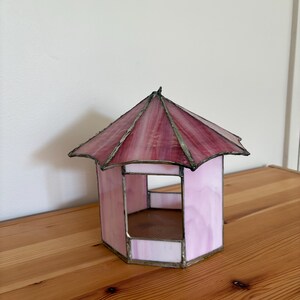 May include: A stained glass gazebo in shades of pink and red, with a hexagonal base and a peaked roof. The glass is framed with a metallic border. The decorative piece sits on a wooden surface.