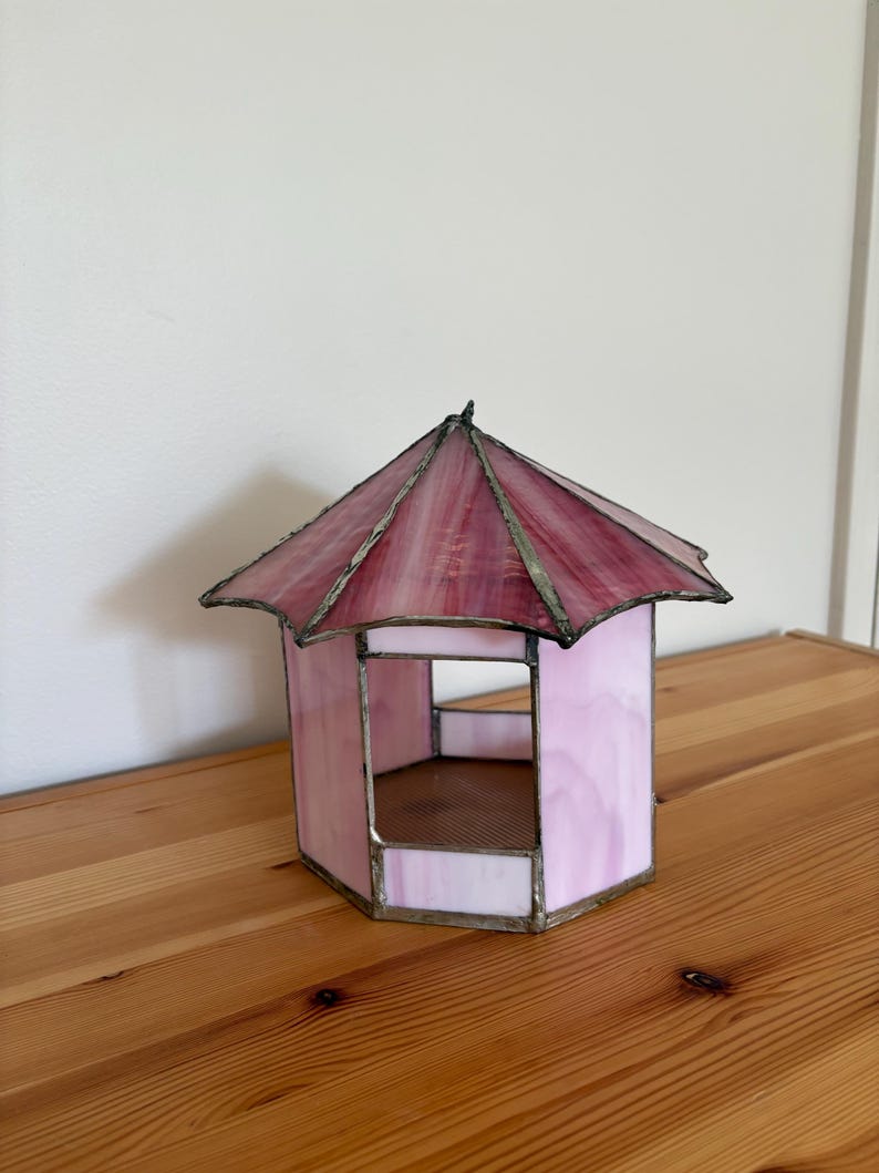May include: A stained glass gazebo in shades of pink and red, with a hexagonal base and a peaked roof. The glass is framed with a metallic border. The decorative piece sits on a wooden surface.