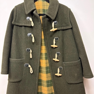 May include: Dark olive green duffel coat with wooden toggles and a plaid lining in yellow and green. The coat has two front pockets and a hood. The coat is hanging on a black hanger.