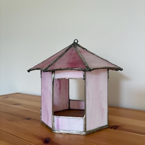 May include: A small, hexagonal stained glass gazebo in shades of pink and purple. The roof and frame are a darker pink, while the walls are a lighter, translucent pink. The glass is held together with a silver-colored metal.