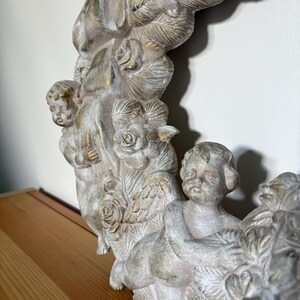 Op de afbeelding: Een gedetailleerde, gebroken witte decoratieve lijst met cherubijnachtige figuren en rozen. De lijst heeft een verweerde, verouderde uitstraling met gouden accenten. Het ontwerp bevat meerdere cherubs en rozen details, wat een klassieke esthetiek cre&euml;ert.