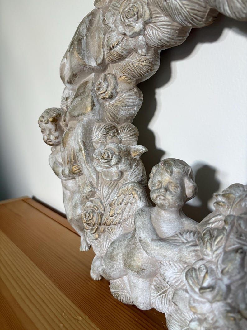 Op de afbeelding: Een gedetailleerde, gebroken witte decoratieve lijst met cherubijnachtige figuren en rozen. De lijst heeft een verweerde, verouderde uitstraling met gouden accenten. Het ontwerp bevat meerdere cherubs en rozen details, wat een klassieke esthetiek cre&euml;ert.