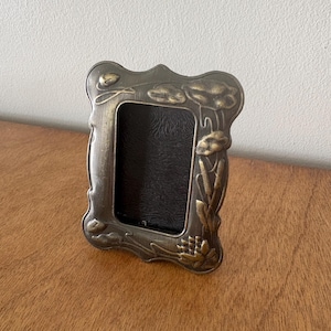 May include: A rectangular picture frame made of antique brass-toned metal with a wavy, scalloped edge. The frame features embossed floral and botanical motifs, including buds and leaves, surrounding a dark, textured rectangular center.