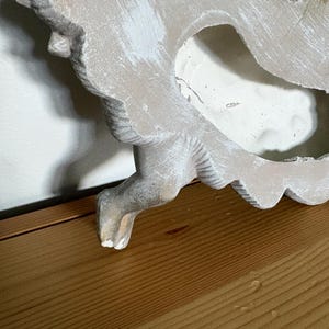Op de afbeelding: Een distressed, gebroken witte decoratieve wandhanger met een gebeeldhouwd, sierlijk ontwerp. Het stuk heeft een verweerde uitstraling met zichtbare penseelstreken van witte verf. Het object is geplaatst op een houten oppervlak.