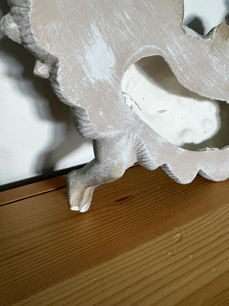 Op de afbeelding: Een distressed, gebroken witte decoratieve wandhanger met een gebeeldhouwd, sierlijk ontwerp. Het stuk heeft een verweerde uitstraling met zichtbare penseelstreken van witte verf. Het object is geplaatst op een houten oppervlak.
