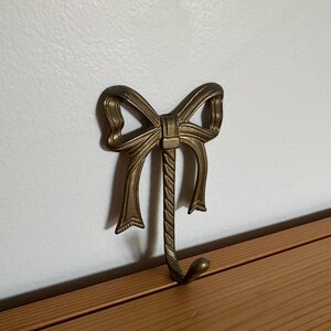 Puede incluir: Un gancho de pared hecho de metal con tono de latón antiguo, que presenta un diseño decorativo de lazo en la parte superior. El gancho tiene una textura trenzada similar a una cuerda que se extiende hacia abajo desde el centro del lazo.
