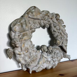 Op de afbeelding: Een decoratieve krans met cherubijnen, rozen en een strik. De krans is geschilderd in een neutraal kleurenpalet met gouden accenten. De cherubijnen zijn in verschillende poses gebeeldhouwd, wat een klassieke en elegante uitstraling geeft.