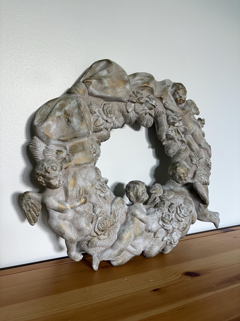 Op de afbeelding: Een decoratieve krans met cherubijnen, rozen en een strik. De krans is geschilderd in een neutraal kleurenpalet met gouden accenten. De cherubijnen zijn in verschillende poses gebeeldhouwd, wat een klassieke en elegante uitstraling geeft.