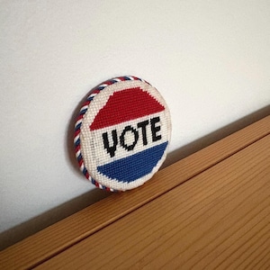 May include: A circular needlepoint patch featuring the word VOTE in black thread. The design includes a red top section, a white middle section with the text, and a blue bottom section. The edge is finished with a twisted red, white, and blue cord.