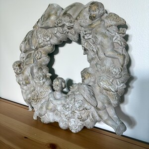 Op de afbeelding: Een gedetailleerde, cirkelvormige kranssculptuur met cherubijnen en rozen. De sculptuur is voornamelijk gebroken wit met gouden accenten, wat een vintage esthetiek cre&euml;ert. De krans is gemaakt van een duurzaam materiaal en is geschikt voor binnenshuis.