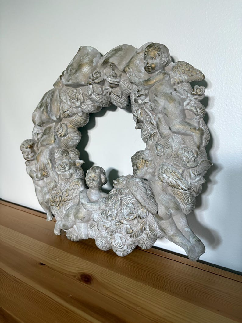 Op de afbeelding: Een gedetailleerde, cirkelvormige kranssculptuur met cherubijnen en rozen. De sculptuur is voornamelijk gebroken wit met gouden accenten, wat een vintage esthetiek cre&euml;ert. De krans is gemaakt van een duurzaam materiaal en is geschikt voor binnenshuis.