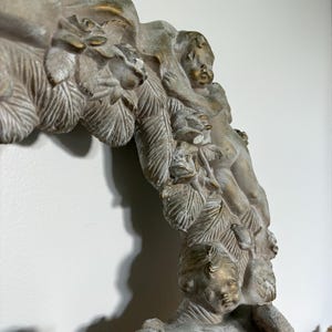 Op de afbeelding: Close-up van een decoratief, antiek ogend framegedeelte. Het frame heeft gedetailleerde gravures van cherubs, bladeren en bloemmotieven. Het kleurenpalet omvat gedempte tinten grijs, wit en goud, wat een vintage esthetiek cre&euml;ert.