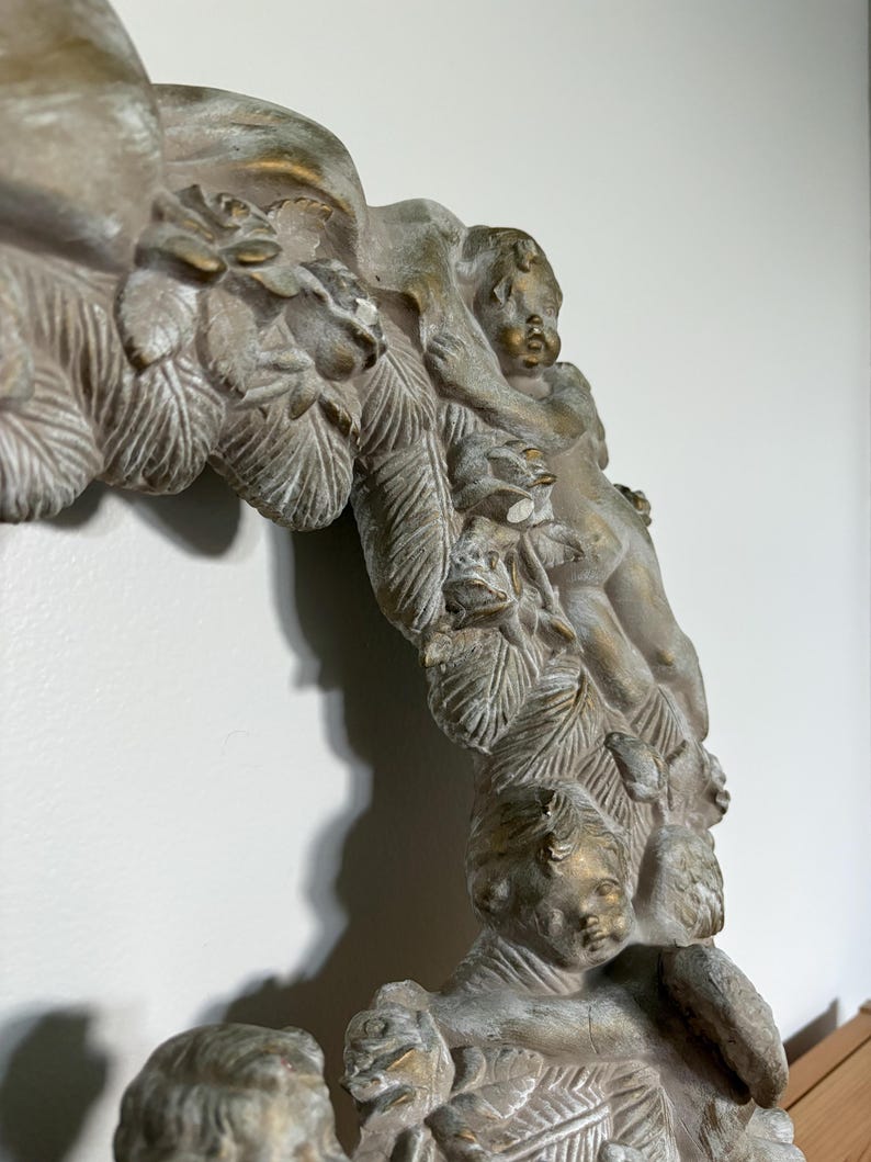 Op de afbeelding: Close-up van een decoratief, antiek ogend framegedeelte. Het frame heeft gedetailleerde gravures van cherubs, bladeren en bloemmotieven. Het kleurenpalet omvat gedempte tinten grijs, wit en goud, wat een vintage esthetiek cre&euml;ert.