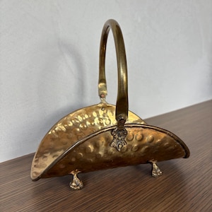 May include: A vintage brass basket with a hammered texture, arched handle, and decorative feet. The basket has a semi-circular shape and is likely used for decorative purposes or storage.