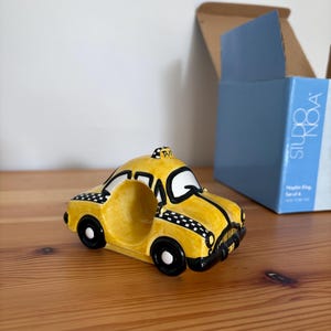 May include: A yellow ceramic taxi-shaped napkin ring with black and white checkered accents. The napkin ring has black wheels and a black front grill. The word "TAXI" is on top of the taxi. A blue box is in the background.