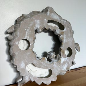 Op de afbeelding: Een verweerde, gebroken witte en grijze decoratieve krans met een centrale opening. De krans heeft gebeeldhouwde details, waaronder een kleine cherubijnfiguur. Het stuk heeft een rustieke, verouderde uitstraling, met zichtbare penseelstreken.