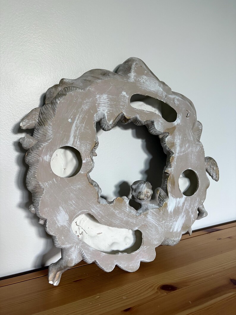 Op de afbeelding: Een verweerde, gebroken witte en grijze decoratieve krans met een centrale opening. De krans heeft gebeeldhouwde details, waaronder een kleine cherubijnfiguur. Het stuk heeft een rustieke, verouderde uitstraling, met zichtbare penseelstreken.