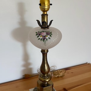 May include: Vintage table lamp with a brass base and a frosted glass shade. The shade is decorated with a hand-painted floral design in pink, purple, and green. The lamp has a square marble base and a gold-colored bulb socket.