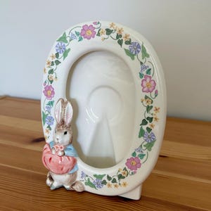 Puede incluir: Marco de fotos ovalado de cerámica con una figura de conejito. El marco es blanco con un borde floral de flores rosas, moradas y amarillas. El conejito sostiene una cesta rosa y lleva una camisa azul.