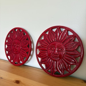 Może przedstawiać: Dwa czerwone, ceramiczne podstawki w kształcie słońca. Każda podstawka ma uśmiechniętą twarz słońca pośrodku, otoczoną stylizowanymi promieniami słonecznymi. Podstawki są okrągłe i mają błyszczące wykończenie.